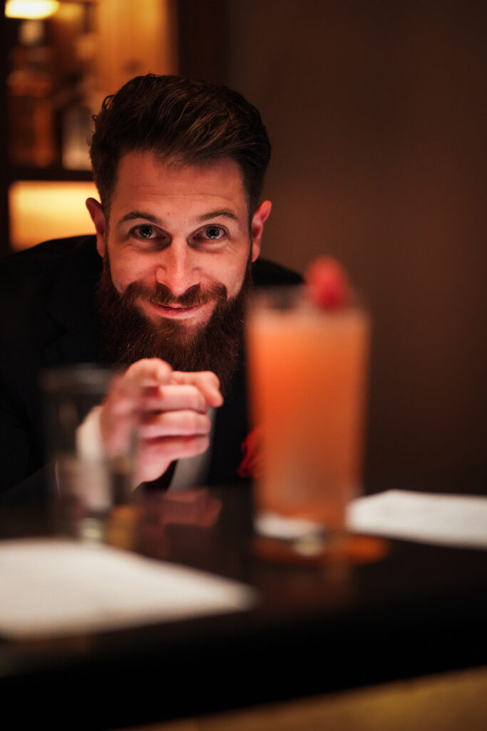Kevin Rigault presenting a cocktail across a bar counter inside a dimly lit hospitality setting. Warm atmosphere focused on guest experience and cocktail service.